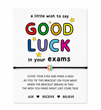 A white rectangular card with colorful text reading 'a little wish to say GOOD LUCK in your exams' and 'ASK RECEIVE BELIEVE' at the bottom. A thin black bracelet with a small rainbow-colored center detail is shown across the middle of the card.