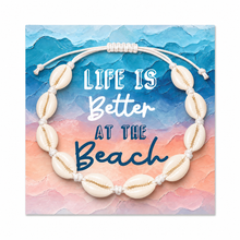 A shell-beaded anklet with white cowrie shells and white rope knots, displayed on a square card with the phrase “LIFE IS Better AT THE Beach” over a blue and peach watercolour beach scene.