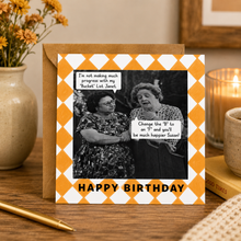 A square birthday card with a yellow and white diamond pattern border, featuring a black-and-white photo of two women with text bubbles and the words 'HAPPY BIRTHDAY' at the bottom. The card is displayed on a wooden table next to a brown envelope, with a candle, flowers, and a pen in the background.