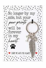 A silver-toned keychain with a feather charm and paw print charms, presented on a white memorial card with black polka dots and a pet loss quote.