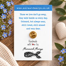 A small gold pin with two interlocking mermaid tails is displayed on a white card with blue text, surrounded by blue flowers, green leaves, a woven textile, and a decorative stone on a wooden surface. The card shows the website mermaidmarys.