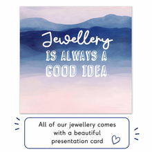 A square presentation card with a watercolor mountain landscape in blue, purple, and pink tones, with the phrase 'Jewellery IS ALWAYS A GOOD IDEA' in white lettering. Below the card is a white banner that reads, 'All of our jewellery comes with a beautiful presentation card' with a small heart icon.