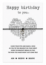 A square greeting card with a heart-shaped word cloud spelling 'Happy Birthday' in black text on a white background. A thin black cotton bracelet with a small silver heart charm is laid across the centre. Birthday card filler.