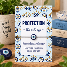 Evil eye bracelet. A square card or box with a blue and white evil eye pattern, tied with a black cord and a blue evil eye bead. The front reads “PROTECTION - The Evil Eye -” and “Peace & Positive Energy Let your intuition guide the way.