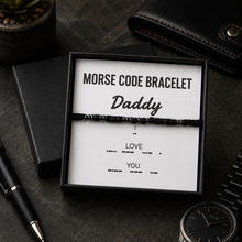A black beaded Morse code bracelet presented in a black gift box with a white insert that reads 'MORSE CODE BRACELET Daddy' and 'LOVE YOU' with Morse code. The box is styled on a dark textured surface with a pen, wallet, and watch nearby.