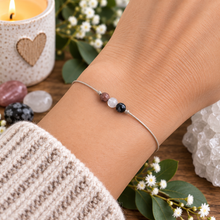 A delicate silver wire bracelet with three small crystal beads (Rhodonite, Moonstone and snowflake Obsidian) worn on a person's wrist, styled with crystals, a candle, and white flowers.