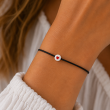 A close-up of a person's wrist wearing a thin black cord bracelet with a small white circular charm featuring a red heart.