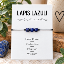 A lapis lazuli beaded bracelet on a navy cord, displayed on a white card labeled 'LAPIS LAZULI' with the brand 'crystals by Mermaid Marys' and spiritual meanings listed below. The card is styled with a blue stone, green leaves, and a ceramic mug in the background.