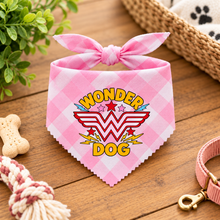 A pink gingham dog bandana with a bow, featuring a 'Wonder Dog' graphic, styled on a wooden tabletop with pet accessories. Wonder dog bandana in pink & white gingham print. Superhero dog bandana. Funny slogan dog scarf neck tie