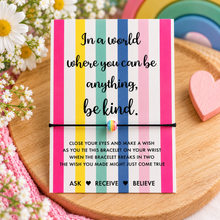 Be kind wish string bracelet. A colourful striped card with a black cord bracelet with a rainbow bead charm wrapped around it. The card reads, 'In a world where you can be anything, be kind.