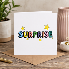 Scratch off panel surprise card. A white greeting card with the word 'SURPRISE' in colorful outlined letters and small yellow stars, standing on a brown envelope on a wooden desk with a plant and pink vase in the background.
