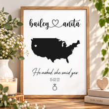 A framed personalized art print featuring a black silhouette map of the United States with a heart over California, the names 'bailey' and 'anita' at the top, and 'He asked, she said yes' with a date and ring illustration below. The print is displayed in a rustic wooden frame on a tabletop, styled with flowers, a candle, books, and greenery.