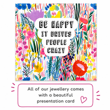 A square floral presentation card with the text 'BE HAPPY IT DRIVES PEOPLE CRAZY' in a white speech-bubble shape, shown above a pink-bordered note that says 'All of our jewellery comes with a beautiful presentation card.'