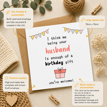 A square white greeting card with a brown kraft envelope, styled on a beige fabric background with decorative dried flowers, twine, and wooden hearts. The card reads, 'I think me being your husband is enough of a birthday gift you're welcome!' with a small pink gift box illustration.