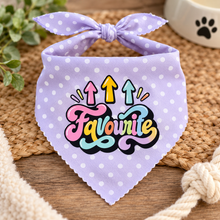 A lavender pet bandana with white polka dots and a bow, featuring a colorful 'Favourite' graphic with three upward arrows. The bandana is styled on a woven mat with a wooden surface, a white rope, and a small plant in the background.