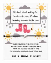 A black cotton cord bracelet with a silver star charm displayed on a white printed card. The card features a whimsical illustration of pink rain boots, yellow suns, gray clouds, and a quote about learning to dance in the rain, with additional text below suggesting a wish that may come true when the bracelet breaks.