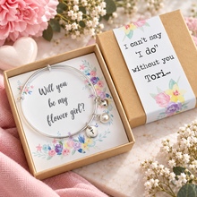 Flower girl proposal charm bangle . A silver bangle bracelet with a small initial 'T' charm and a flower and pearl charms, presented in a floral gift box next to its kraft paper sleeve. The box contains a card that reads 'Will you be my flower girl?' and the sleeve reads 'I can't say 'I do' without you, and the recipients name. Flower girl gift - weddings favours 