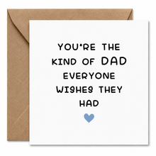 A white square greeting card with black hand-lettered text reading 'YOU'RE THE KIND OF DAD EVERYONE WISHES THEY HAD' and a small light blue heart, shown with a brown kraft envelope behind it on a white background.