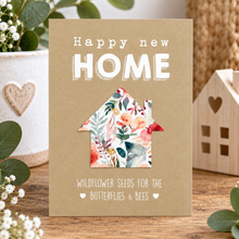 A plantable seed paper gift. A card with the words 'Happy new HOME' and 'WILDFLOWER SEEDS FOR THE BUTTERFLIES & BEES' displayed on a wooden tabletop, surrounded by decorative plants and a small wooden house.