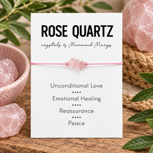 A pink cord bracelet with three rose quartz beads displayed on a white card that reads 'ROSE QUARTZ crystals by Mermaid Marys' and lists benefits. The card is styled on a wooden surface with pink crystal clusters and green leaves around it.