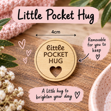 A round wooden token engraved with 'little POCKET HUG' and a heart cutout, displayed on a wooden surface with decorative flowers, a pink knitted item, and text overlays showing dimensions and product details.
