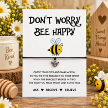 Bee wish string bracelet. A bee-themed charm bracelet presented on a white card with printed instructions and the phrase 'DON'T WORRY BEE HAPPY.' The card is styled on a wooden surface with flowers, a honey pot, and a wooden house in the background.