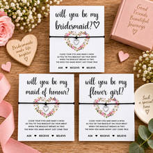 Three white bridal bracelets with message cards. The cards read 'will you be my bridesmaid?', 'will you be my maid of honour?', and 'will you be my flower girl?' with floral heart graphics. Wedding proposal wish string bracelets.