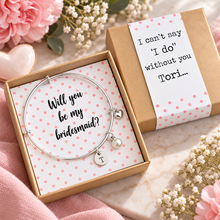 Personalised Bridesmaid proposal bangle. A silver bangle bracelet with a small heart charm and a letter 'T' initial pendant is displayed in an open kraft gift box with pink polka dots. The box contains a white card that reads 'Will you be my bridesmaid?'. Bridesmaid gift. 