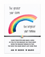 Rainbow wish bracelet. A square white card with a colourful watercolour rainbow and the phrases 'The greater your storm the brighter your rainbow.' A thin black bracelet with a small striped bead is shown across the center.