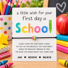 First day at school wish string bracelet. A school-themed first-day-of-school bracelet card on a wooden desk surrounded by school supplies. The white card features a colorful 'School' word design with a small striped beaded black cord bracelet attached.
