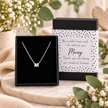 A silver initial necklace with the letter 'M' displayed in an open black gift box next to its black and white polka-dot presentation box with a custom thank you message, styled on a marble tray with flowers.