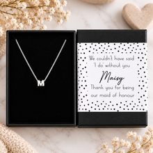 A silver initial necklace with the letter 'M' displayed inside an open black gift box next to its printed card. The card reads, 'We couldn't have said 'I do' without you Maizy Thank you for being our maid of honour'.
