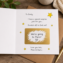 An open handmade surprise card with printed text and a gold foil rectangle on a white page, placed on a brown envelope over a wooden surface with a plant leaf, stone, and pen visible around it. Scratch off card
