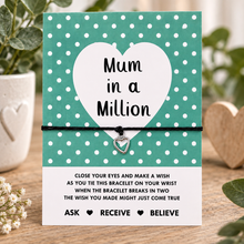 A square greeting display card with a teal and white polka dot design, featuring a large white heart in the centre with the text 'Mum in a Million.' A black cotton cord bracelet with a small silver heart charm is tied across the middle. Mother’s Day gift for mum.