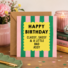 A square greeting card with pink and green vertical stripes and a yellow scalloped center panel reading 'HAPPY BIRTHDAY CLASSY, SASSY & A LITTLE BAD ASSY' in black text, shown with a brown envelope on a tabletop with flowers, books, and a candle in the background.