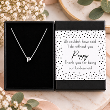 A silver initial necklace with the letter 'P' displayed inside an open black gift box next to a printed message card that reads, 'We couldn't have said 'I do' without you Poppy Thank you for being our bridesmaid.' The boxes are styled on a marble surface with dried flowers and green leaves.