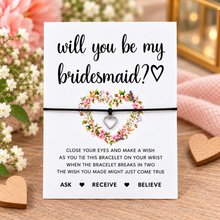 A 'Will You Be My Bridesmaid?' Bracelet set. The white card features black text, a floral heart design, and instructions for the wish, with a black cord bracelet and silver heart charm wrapped around it. Bridesmaid proposal wish string bracelet. 