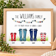 A framed personalised family print featuring the text 'THE WILLIAMS FAMILY' and 'JUST THE RIGHT AMOUNT OF CHAOS & LOVE!' above an illustration of four rain boots labeled 'POPPY', 'DADDY', and 'TILLY'. The print is shown in a light wood frame