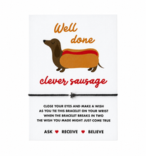 Clever sausage wish string bracelet. A white congratulations card with a dachshund in a hot dog illustration and the words 'Well done clever sausage,' tied with a black bracelet with a silver seat charm. Congratulations gift. Gift for new job or passing exams 