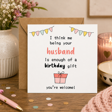 Wife birthday card. A square white birthday card with a brown kraft envelope, placed on a wooden table in a styled home setting. The card reads, 'I think me being your husband is enough of a birthday gift you're welcome!' with pastel bunting and a small gift box illustration.