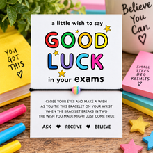A white rectangular card with colorful text reading 'a little wish to say GOOD LUCK in your exams,' tied with a small striped bracelet, surrounded by pastel pens, star-shaped objects, and motivational notes on a wooden surface.