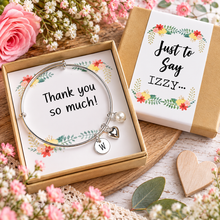 A silver expandable bracelet with a pearl charm, heart charm, and initial 'W' charm, presented in a kraft gift box with a floral insert reading 'Thank you so much!'. Personalised silver thank you charm bangle. Thank you gift 