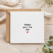 A square white greeting card with the printed phrase 'Happy you + me day' in black lowercase letters and a small red heart, shown with a brown kraft envelope on a light wood surface with soft decorative props.