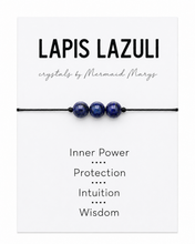A Lapis Lazuli crystal bracelet with three polished deep blue beads on a black cord, presented on a white card labeled 'LAPIS LAZULI' with the brand 'crystals by Mermaid Marys' and spiritual meanings listed below.