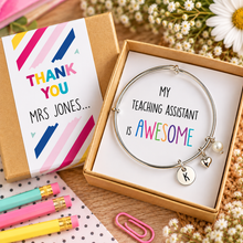Personalise teaching assistant bangle with 3 charms. Silver charm bracelet presented in a kraft gift box with a white insert that reads 'MY TEACHING ASSISTANT IS AWESOME'. The box lid beside it has a colorful 'THANK YOU MRS JONES. TA gift