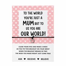 A heart-themed charm bracelet presented on its original packaging card. The card is pink with a pattern of small white hearts at the top, and a white panel in the centre with the text 'TO THE WORLD YOU'RE JUST A MUM BUT TO US YOU ARE OUR WORLD!'. Black cotton cord bracelet with small silver pink heart charm. 