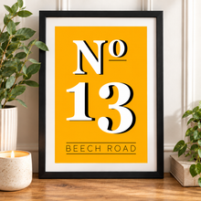 A framed typographic art print reading “No 13 Beech Road” in bold white lettering on a mustard yellow background, displayed on a wooden surface with plants and a candle around it.