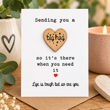 A square white greeting card with black printed text reading 'Sending you a big hug so it's there when you need it' and 'Life is tough but so are you,' displayed on a wooden surface with a small wooden heart, a lit candle, greenery, and a ceramic vase in the background.