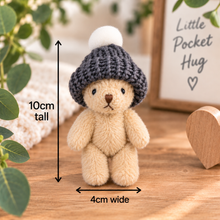 A small plush teddy bear wearing a dark gray knit beanie with a white pom-pom, shown on a wooden surface with measurement arrows indicating 10cm tall and 4cm wide. Decorative leaves, a framed sign reading 'Little Pocket Hug,' and a wooden heart are in the background.