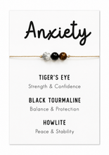 A beaded anxiety bracelet displayed on a white card. The bracelet consists of three beads: a silver marbled bead, a black tourmaline bead, and a brown tiger's eye bead, strung on a thin gold cord.